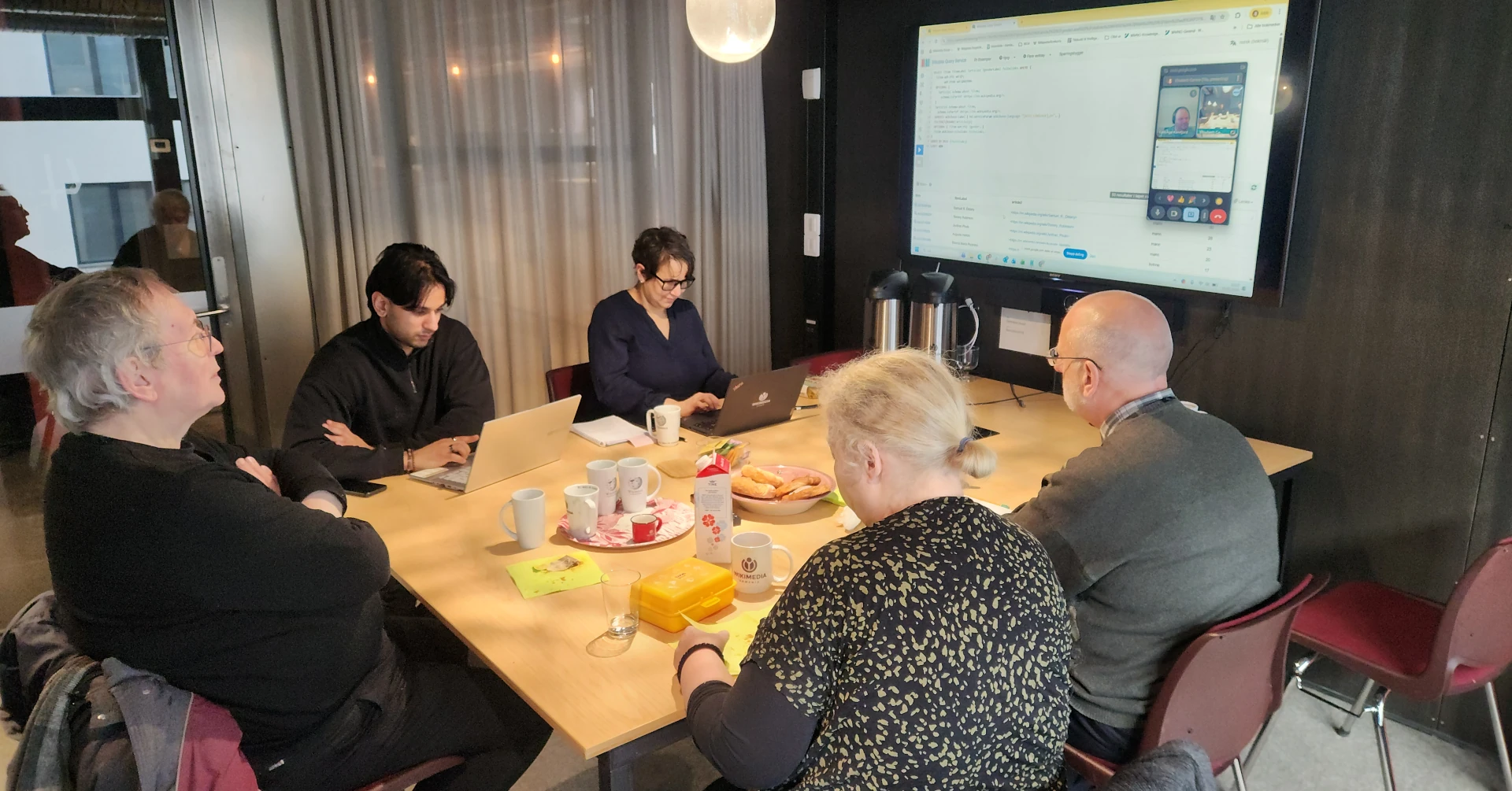 Fem personer på et møterom rundt et bord med en sjette person på skjerm. Arrangementet "Wikilunsj" på Wikimedia Norge sine kontorer 5. februar 2026.